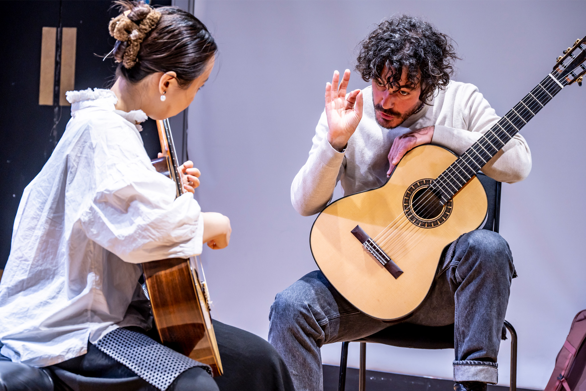 A man wearing a white shirt, holding his guitar, teaching a male student, holding and strumming his guitar, while listening.